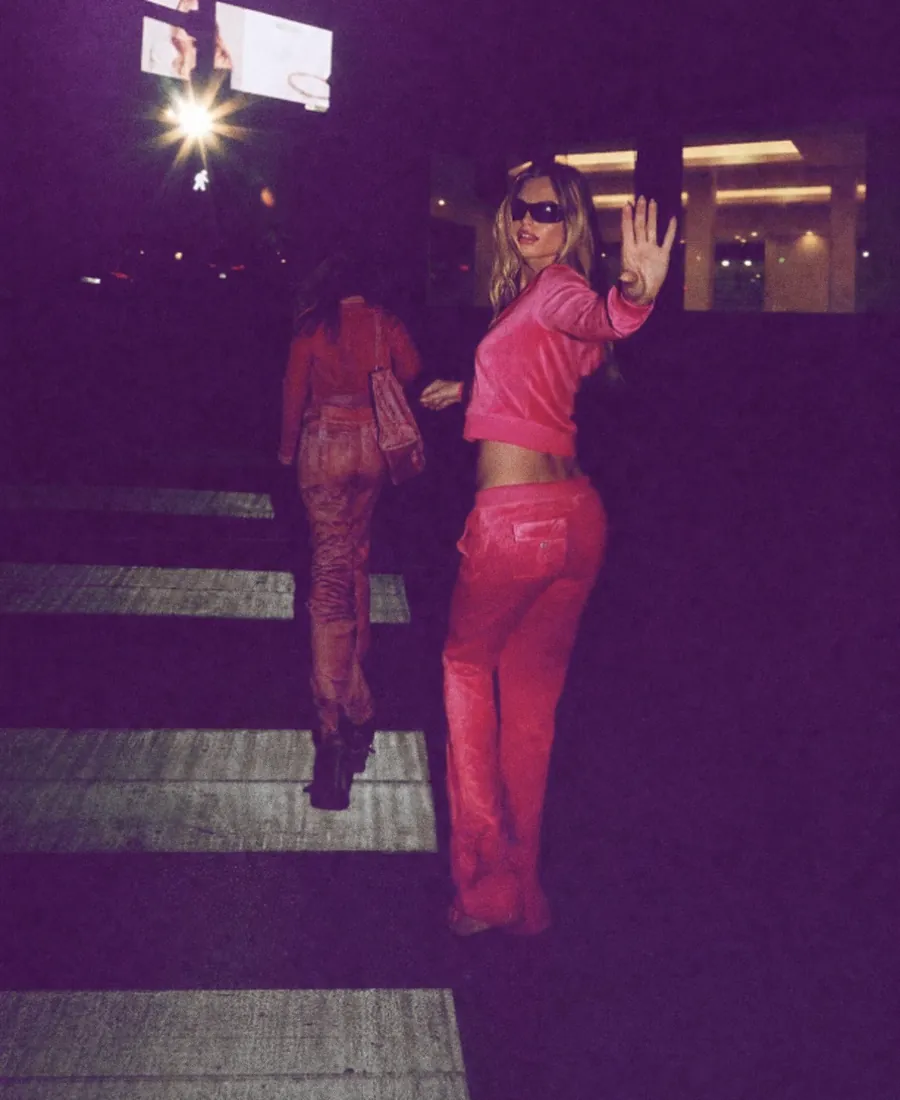 Due ragazze in tuta rosa attraversano la strada di notte, una saluta verso la macchina fotografica